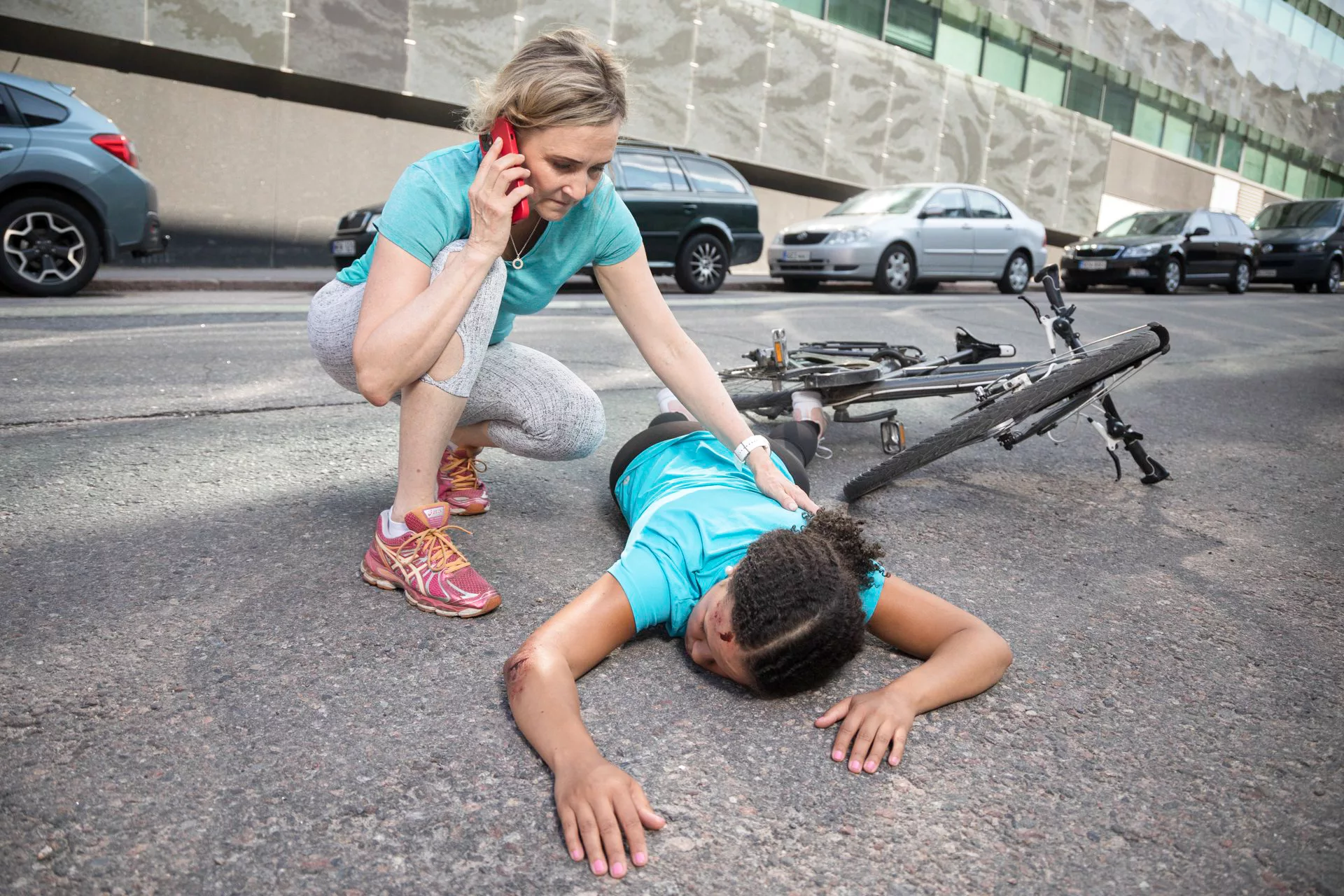 En kvinna har ramlat av sin cykel och ligger medvetslös på gatan. En annan kvinna ringer efter hjälp på telefon.